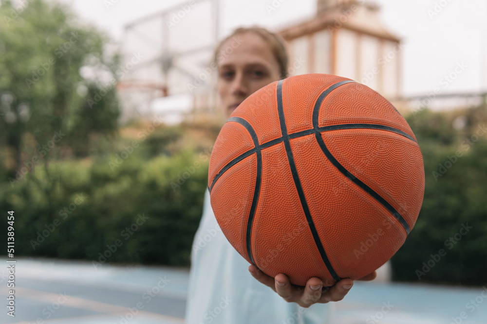 American blonde girl offering a basketball on the court. Close-up image of a sporty blurry woman displaying an orange ball challenging her opponent to start a street basketball game. Sport concept American blonde girl offering a basketball on the court. Close-up image of a sporty blurry woman displaying an orange ball challenging her opponent to start a street basketball game. Sport concept