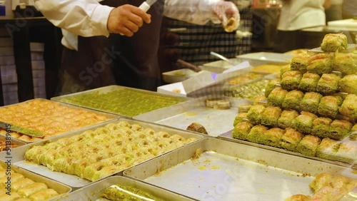 Stacks of baklava at foreground and hands of unrecognizable seller serves customers at traditional turkish delights shop. Showcase of turkish confectionery