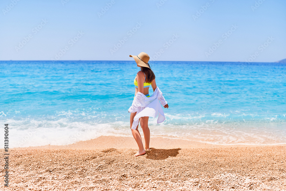 Back of fashionable gorgeous stylish sexy slim beautiful brunette woman in straw hat, bright swimsuit and shirt on sea beach in summer sunny day Back of fashionable gorgeous stylish sexy slim beautiful brunette woman in straw hat, bright swimsuit and shirt on sea beach in summer sunny day