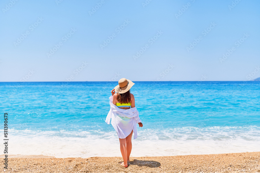 Back of fashionable gorgeous stylish sexy slim beautiful brunette woman in straw hat, bright swimsuit and shirt on sea beach in summer sunny day Back of fashionable gorgeous stylish sexy slim beautiful brunette woman in straw hat, bright swimsuit and shirt on sea beach in summer sunny day