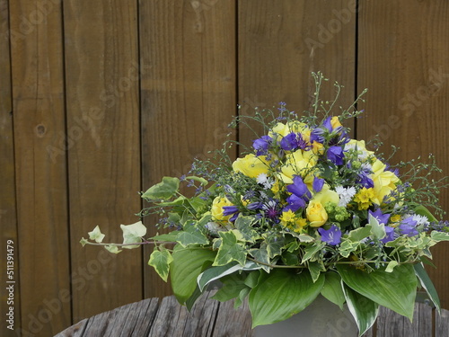 Strauss aus echten Blüten mit blauen Glockenblumen und gelben Rosen, weissen Matrikaria, Leinkraut und Funkienblättern vor Hintergrund aus Holzdielen