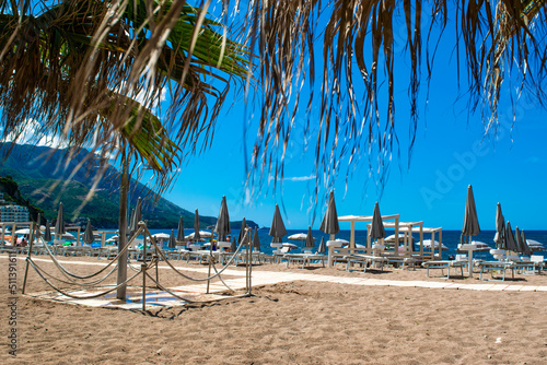 Rows of umbrellas and sun loungers against backdrop of palm leaves on public sundy beach at sunny day. Calm sea and blue sky. Becici, Montenegro. photo