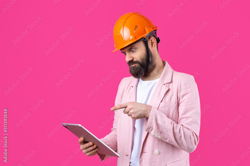 Portrait of young construction engineer wear orange hard hat, in a pink jacket standing on red studio background. A man with a tablet in his hands. Portrait of young construction engineer wear orange hard hat, in a pink jacket standing on red studio background. A man with a tablet in his hands.