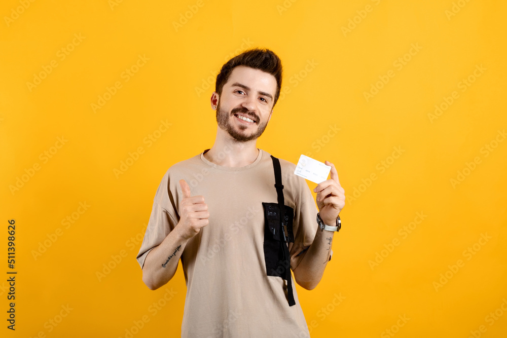 Happy young man wearing beige t-shirt posing isolated over yellow background holding credit bank card showing thumb up. Mock up copy space. Happy young man wearing beige t-shirt posing isolated over yellow background holding credit bank card showing thumb up. Mock up copy space.