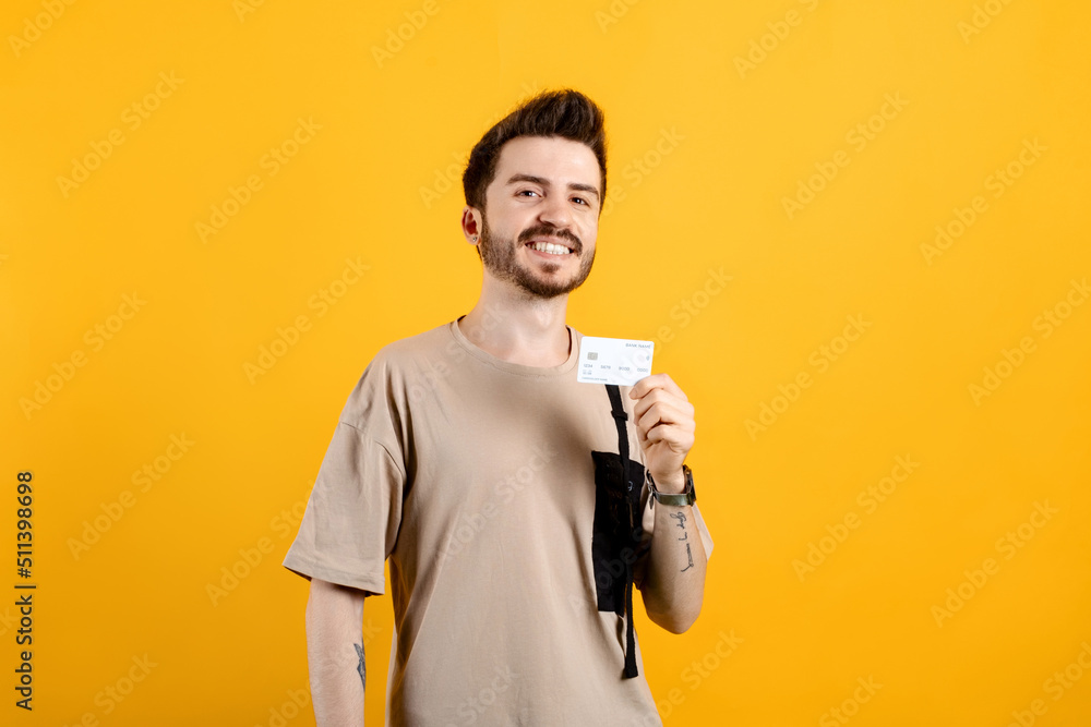 Young man smiling confident wearing beige tee posing isolated over yellow background holding plastic credit or debit card while looking at the camera. Young man smiling confident wearing beige tee posing isolated over yellow background holding plastic credit or debit card while looking at the camera.