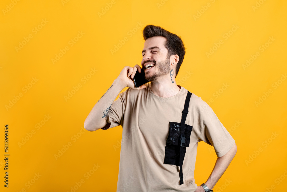 Happy young man wearing beige t-shirt posing isolated over yellow background talking on smart phone and smiling. Receiving good news during phone conversation. Happy young man wearing beige t-shirt posing isolated over yellow background talking on smart phone and smiling. Receiving good news during phone conversation.