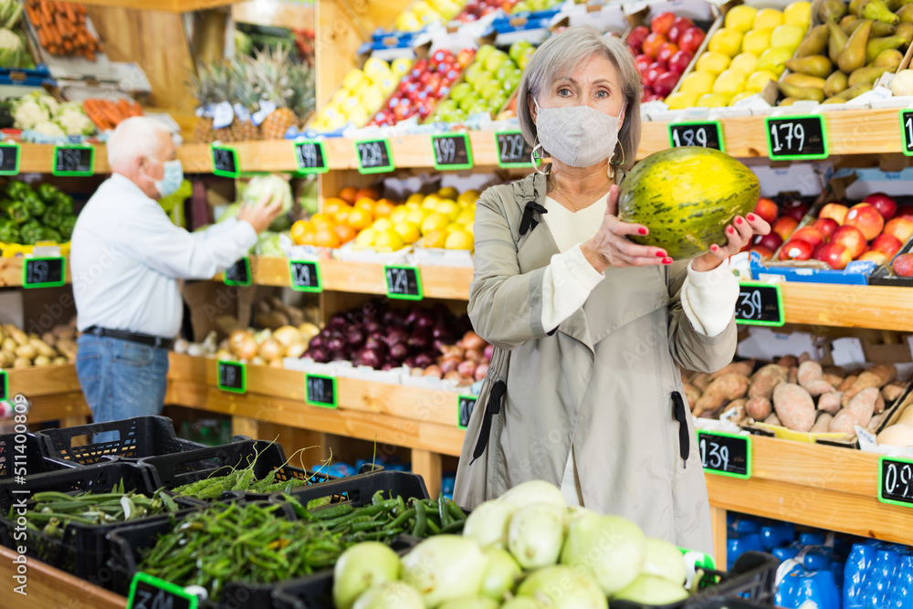 Woman in protective mask selecting watermelon in greengrocer. Man walking in background