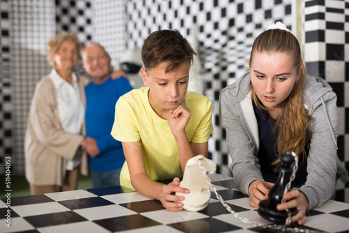 Young girl and boy standing in escape room and resolving chess conundrum. Their grandparents standing behind them and looking at them.