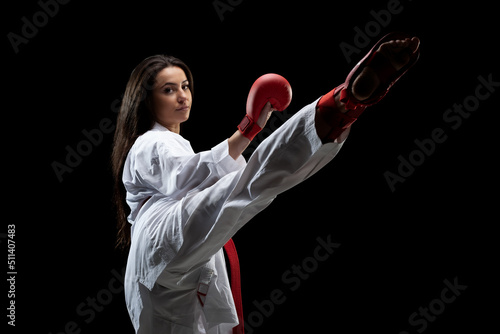 girl exercising karate leg kick wearing kimono and red gloves against black background..