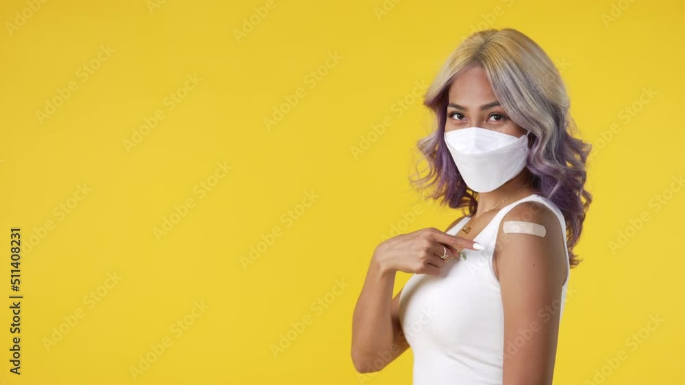 Young Asian woman wearing mask and getting a vaccine on yellow background. Woman showing her arm with bandage after receiving vaccination. Young Asian woman wearing mask and getting a vaccine on yellow background. Woman showing her arm with bandage after receiving vaccination.