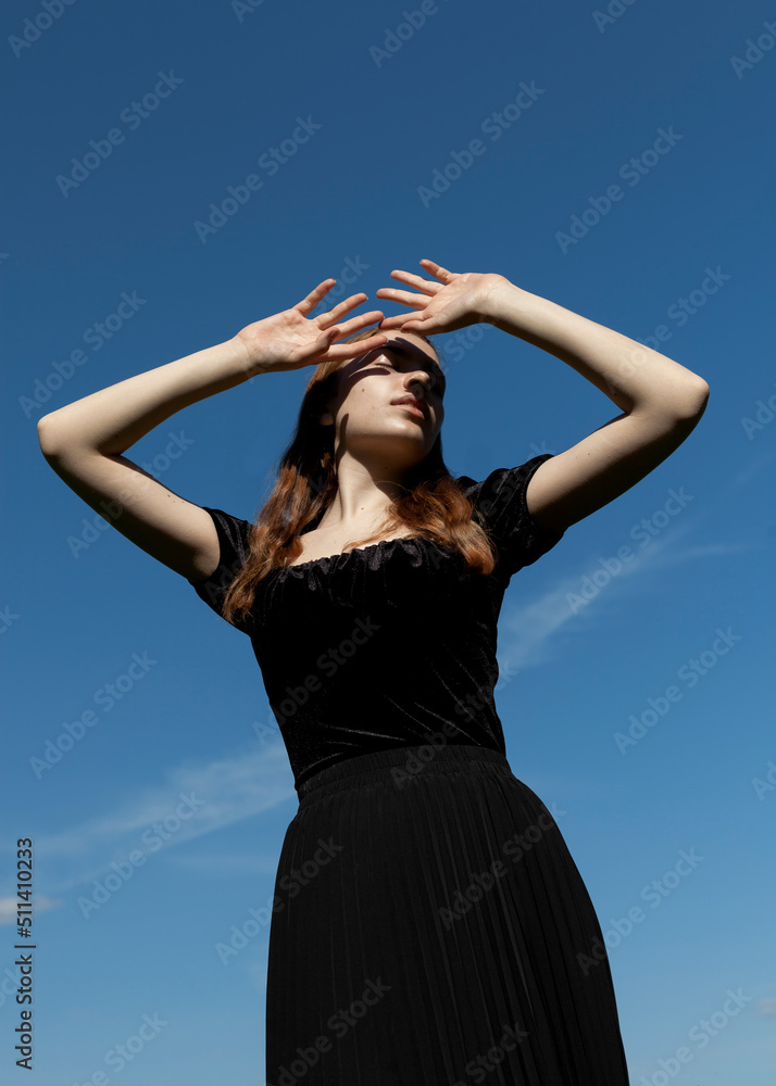 an elegant woman in a black dress stands against the blue sky in sunny weather covering her face with her hands an elegant woman in a black dress stands against the blue sky in sunny weather covering her face with her hands