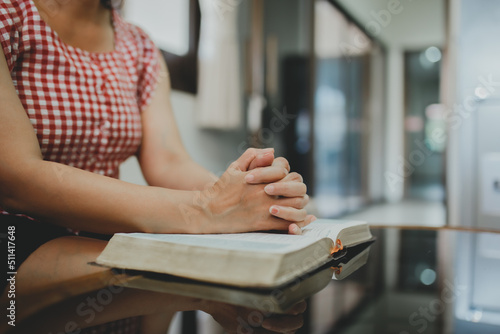 Fotografiet Close up of a woman hands praying on the open holy  bible on a table indoor with the windows light lay warm tone