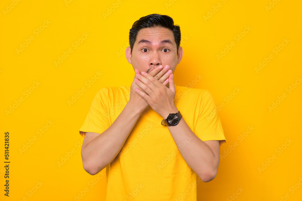 Shocked young Asian man in casual t-shirt covering mouth with hands and looking at camera isolated on yellow background. People lifestyle concept Shocked young Asian man in casual t-shirt covering mouth with hands and looking at camera isolated on yellow background. People lifestyle concept