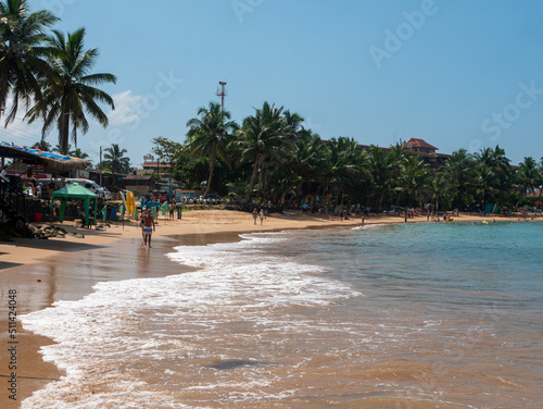 Fototapeta Naklejka Na Ścianę i Meble -  Hikkaduwa, Sri Lanka - March 8, 2022: Beautiful view of Hikkaduwa beach with green palm trees against the blue sky. People walk on the sand along the Indian Ocean