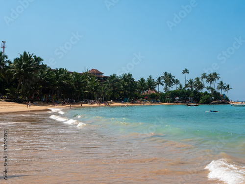 Hikkaduwa, Sri Lanka - March 8, 2022: Beautiful view of Hikkaduwa beach with green palm trees against the blue sky. People relax, sunbathe and swim in the azure water of the Indian Ocean. Copy space