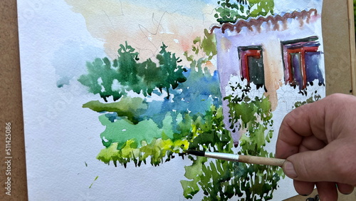 the picture is painted with paints on it depicts a house on the windowsill flowers and around a lot of greenery painting in process of drawing Because some details are underpainted just a white place. photo