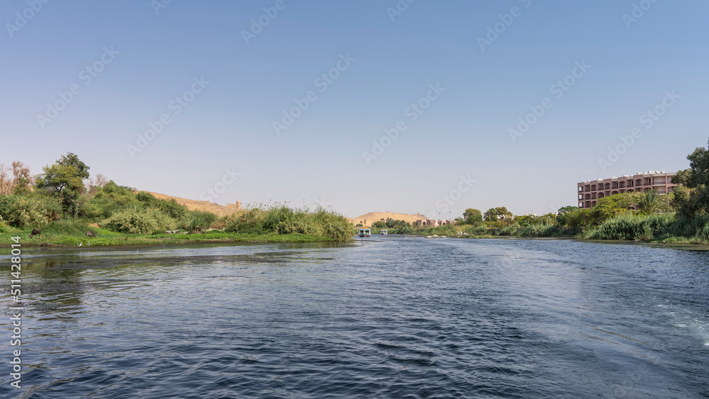 The river flows calmly. There is green vegetation on the banks. A tourist boat is sailing on blue water. Sand dunes against a clear sky. Copy space. Egypt. Nile