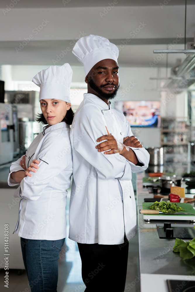 Portrait of gastronomy experts standing back to back inside restaurant professional kitchen while smiling confident at camera. Diverse food industry workers posing for camera.