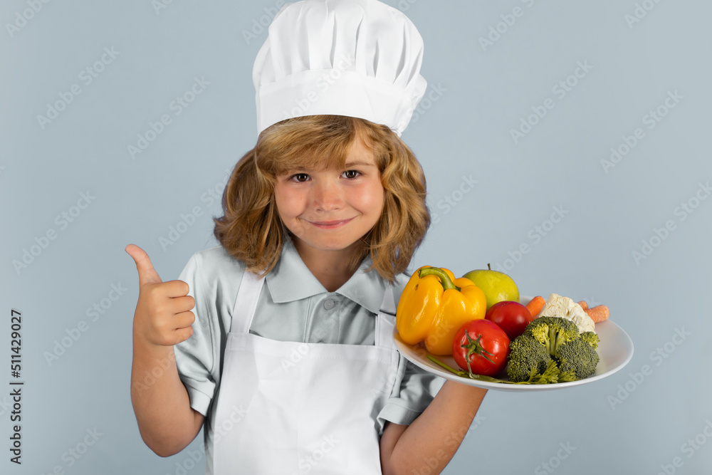 Child wearing cooker uniform and chef hat hold plate with vegetables preparing vegetables on kitchen, studio portrait. Cooking, culinary and kids food concept. Child wearing cooker uniform and chef hat hold plate with vegetables preparing vegetables on kitchen, studio portrait. Cooking, culinary and kids food concept.