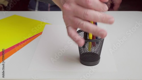 The artist puts colored pencils in a stand. A glass of colored pencils on a white table with drawing paper.
