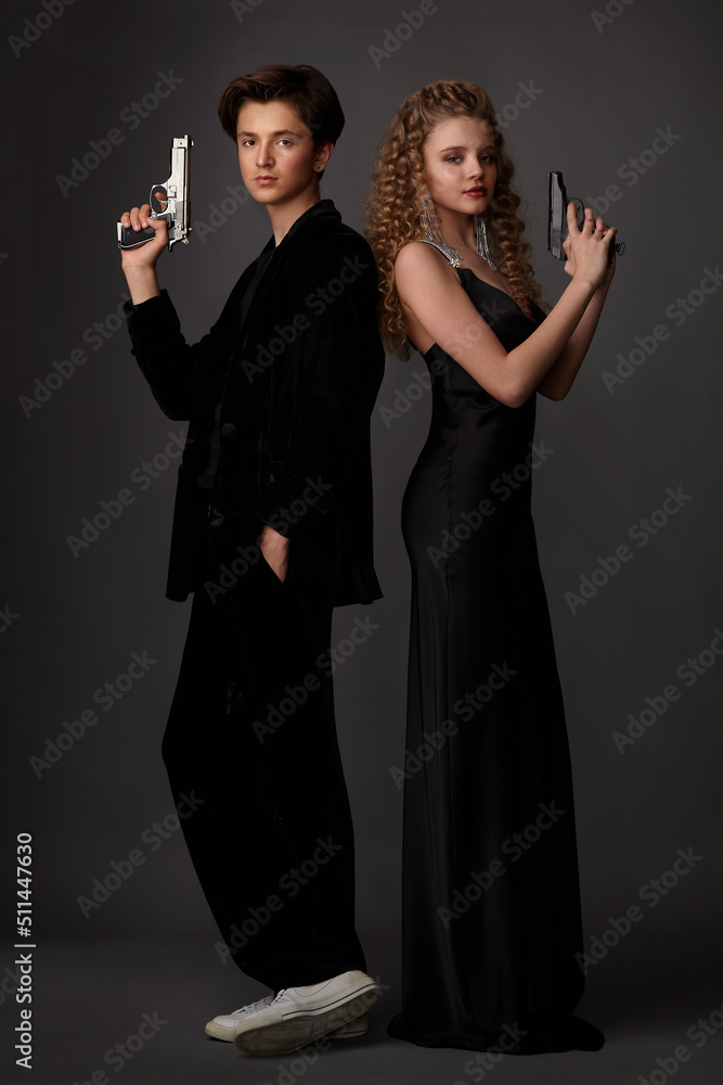 A guy in a black suit and a girl in a black long dress
with pistols in their hands posing in the studio on a gray background A guy in a black suit and a girl in a black long dress
with pistols in their hands posing in the studio on a gray background