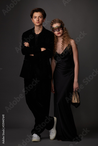 A guy in a black suit and a girl in a black long dress with shoes in their hands posing in the studio on a gray background