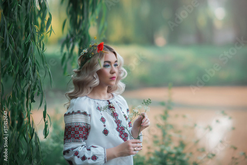 A blonde woman with a flower in her hair sits in tall green grass and looks into the distance. Beautiful blonde with evening make-up and long curls in nature.