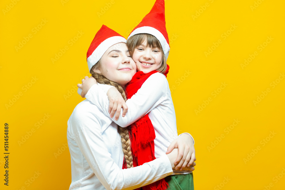 Fototapeta premium Family of Two Positive Caucasian Girls Sisters In Festive Santa Hats Having Fun While Embracing And Toching by Cheeks On Yellow Background.