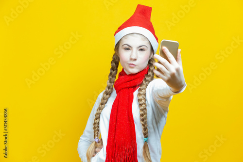 Funny Winsome Caucasian Teenager Holding Cellphone Taking Selfie While Smiling and Having Good Time And Making Faces in Christmas Santa Hat Posing Against Yellow