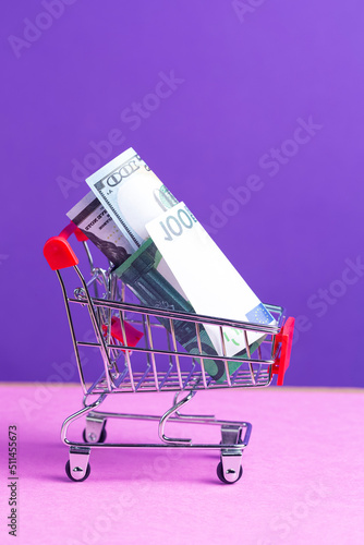 Savings Ideas. Shopping Trolley Cart With US Dollars and Euro Currency Banknotes Over Trendy Purple Violet Background.
