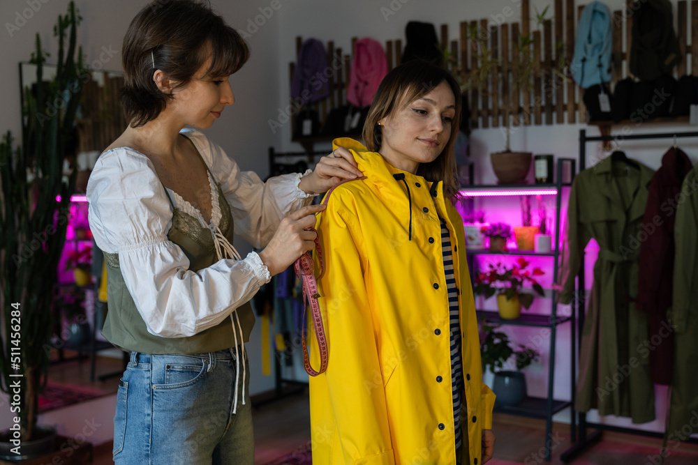 Making measures before tailoring. Fashion designer workshop with female tailor measuring and fitting bespoke rain jacket. Young woman small dressmaking business owner at workplace. Needlework concept 
