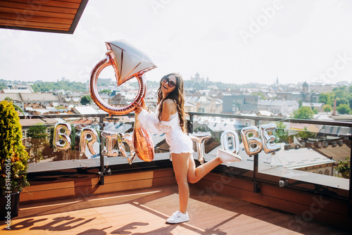 Bachelorette party. Beautiful girl in a white shirt and shorts posing with a balloon in the form of a wedding ring on the roof of the house.