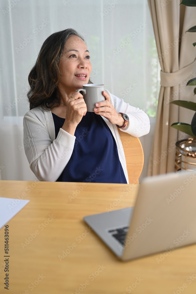 Relaxed middle aged woman holding cup of coffee, daydreaming while looking away. Retirement, technology concept Relaxed middle aged woman holding cup of coffee, daydreaming while looking away. Retirement, technology concept