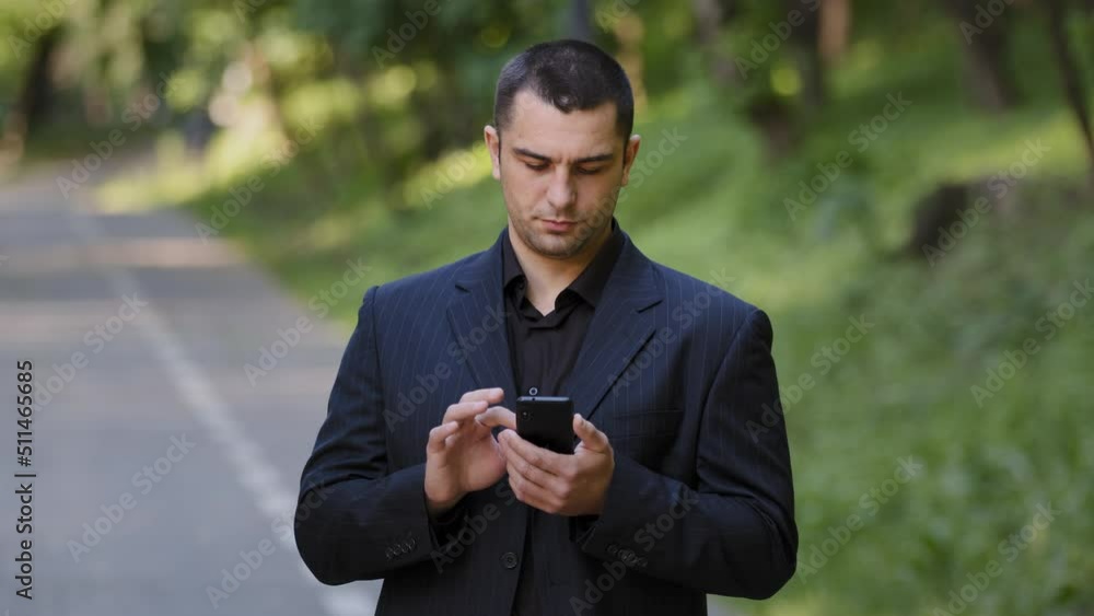 Serious adult caucasian man user stand outdoors in park wears black formal suit hold mobile telephone scrolling slide pages on smartphone screen online information texting message social business chat Serious adult caucasian man user stand outdoors in park wears black formal suit hold mobile telephone scrolling slide pages on smartphone screen online information texting message social business chat