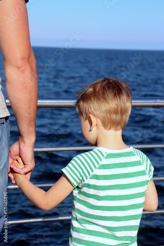 a boy in a striped T-shirt stands by the sea and holds his father's hand rear view selective focus