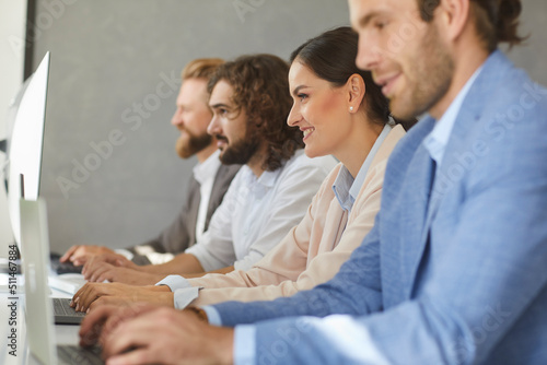 Group of smiling business people using modern software on desktop and laptop computers. Side view of team of happy satisfied employees sitting in row at office table, looking at screens and typing