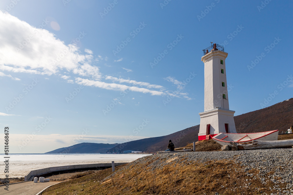 Lighthouse (leading beacon) and a boat on the shore of the sea bay. View from the shore to the ice-covered bay. Nagaev Bay, Sea of Okhotsk. Park Lighthouse in the city of Magadan. Far East of Russia.