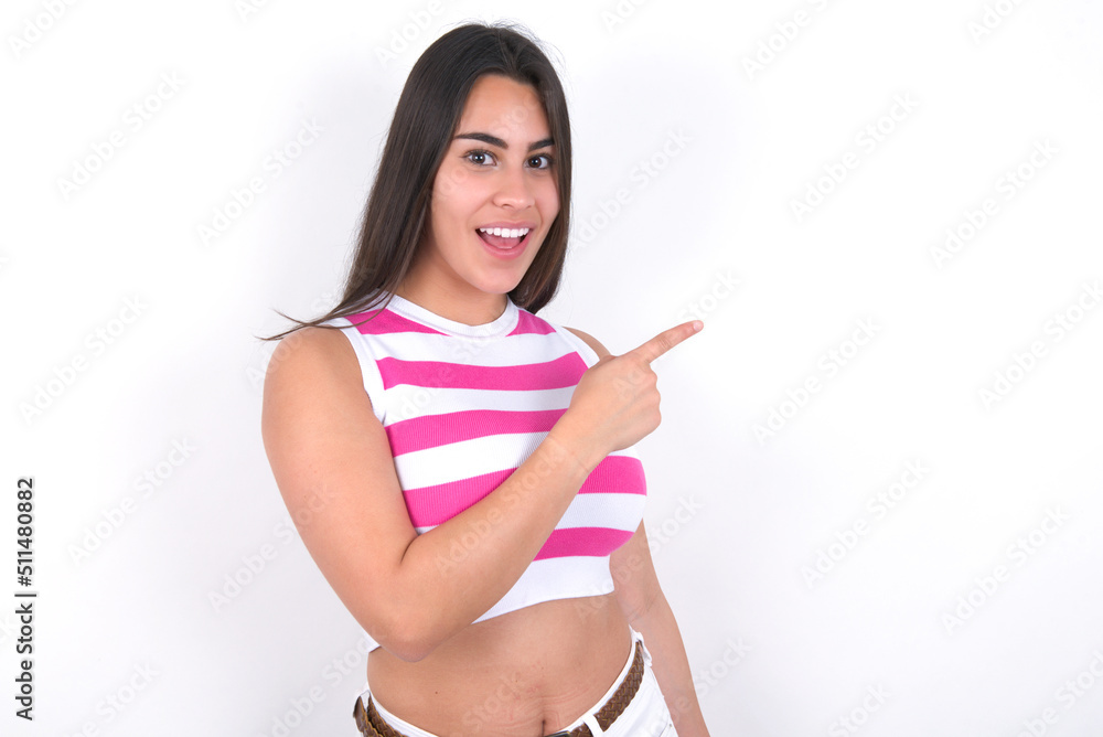 young beautiful brunette woman wearing striped top over white wall pointing away and smiling to you. Look over there! young beautiful brunette woman wearing striped top over white wall pointing away and smiling to you. Look over there!