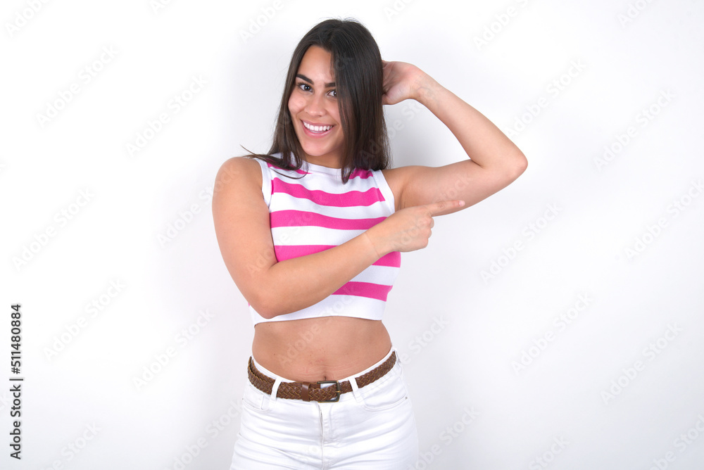 young beautiful brunette woman wearing striped top over white wall feeling positive has amazed expression, indicates something. One hand on head and pointing with other hand. young beautiful brunette woman wearing striped top over white wall feeling positive has amazed expression, indicates something. One hand on head and pointing with other hand.