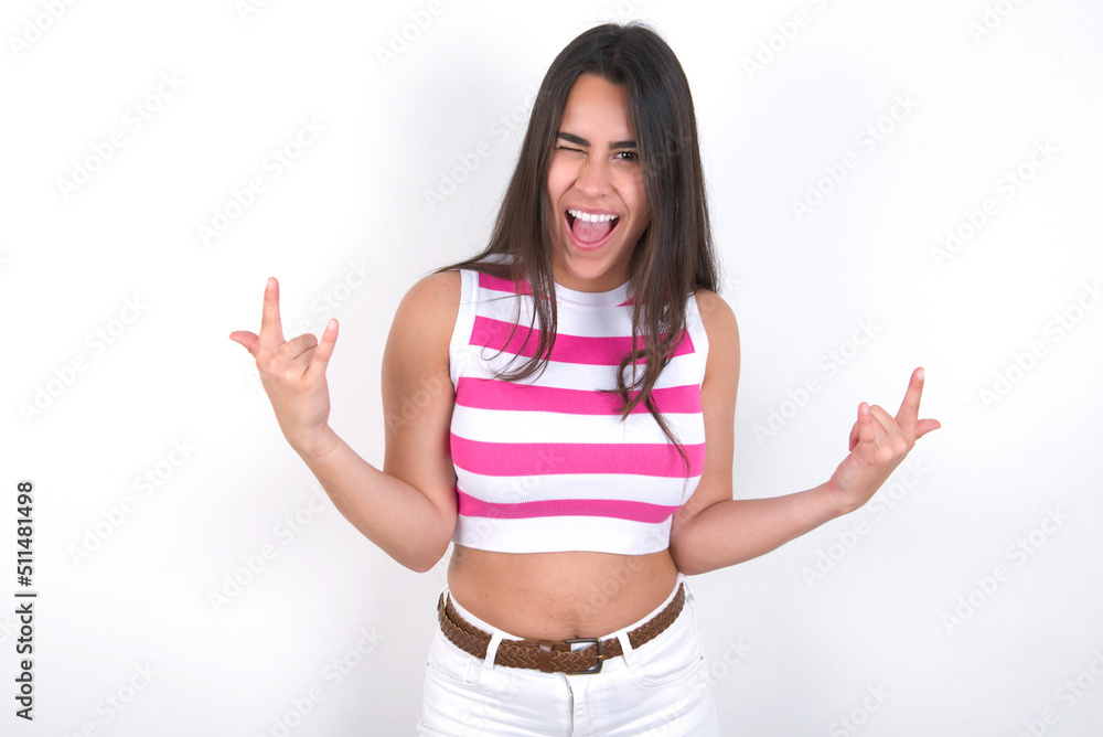 Born to rock this world. Joyful young beautiful brunette woman wearing striped top over white wall screaming out loud and showing with raised arms horns or rock gesture. Born to rock this world. Joyful young beautiful brunette woman wearing striped top over white wall screaming out loud and showing with raised arms horns or rock gesture.