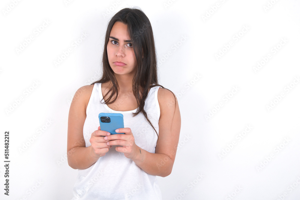 Portrait of a confused Young beautiful brunette woman wearing white top over white wall holding mobile phone and shrugging shoulders and frowning face. Portrait of a confused Young beautiful brunette woman wearing white top over white wall holding mobile phone and shrugging shoulders and frowning face.