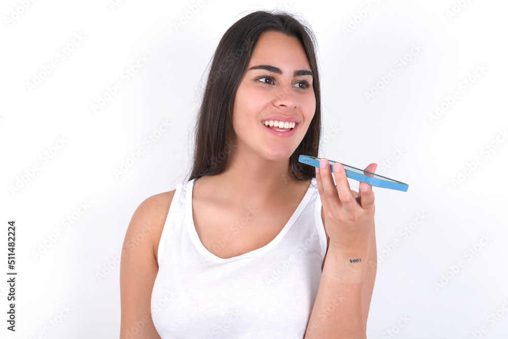 Smiling Young beautiful brunette woman wearing white top over white wall sending voice message on her smart phone. Communication and new technologies concept. Smiling Young beautiful brunette woman wearing white top over white wall sending voice message on her smart phone. Communication and new technologies concept.