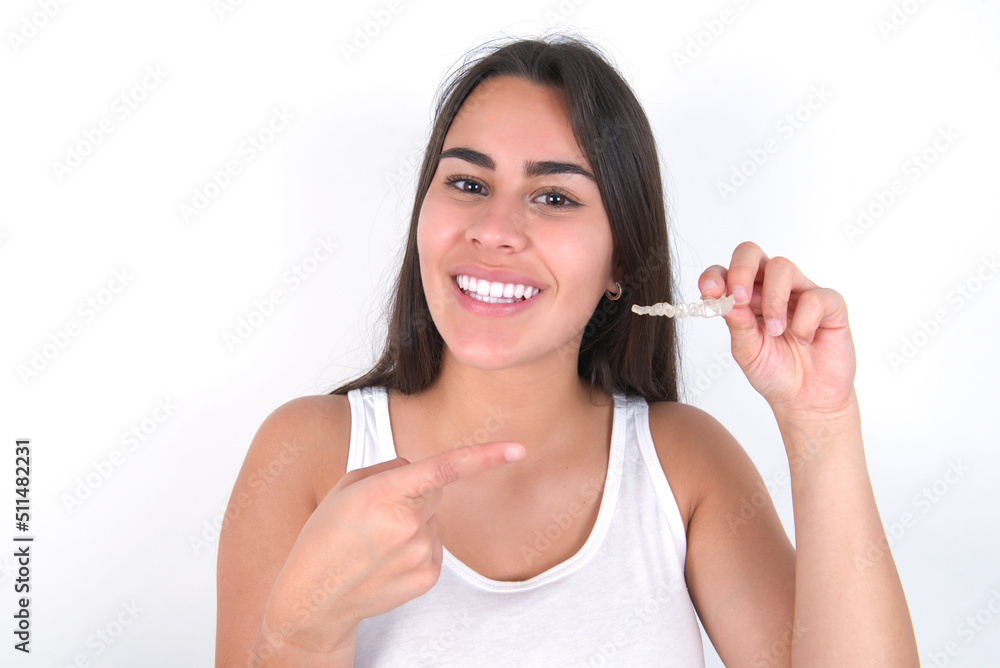 Young beautiful brunette woman wearing white top over white wall holding an invisible aligner and pointing at it. Dental healthcare and confidence concept. Young beautiful brunette woman wearing white top over white wall holding an invisible aligner and pointing at it. Dental healthcare and confidence concept.