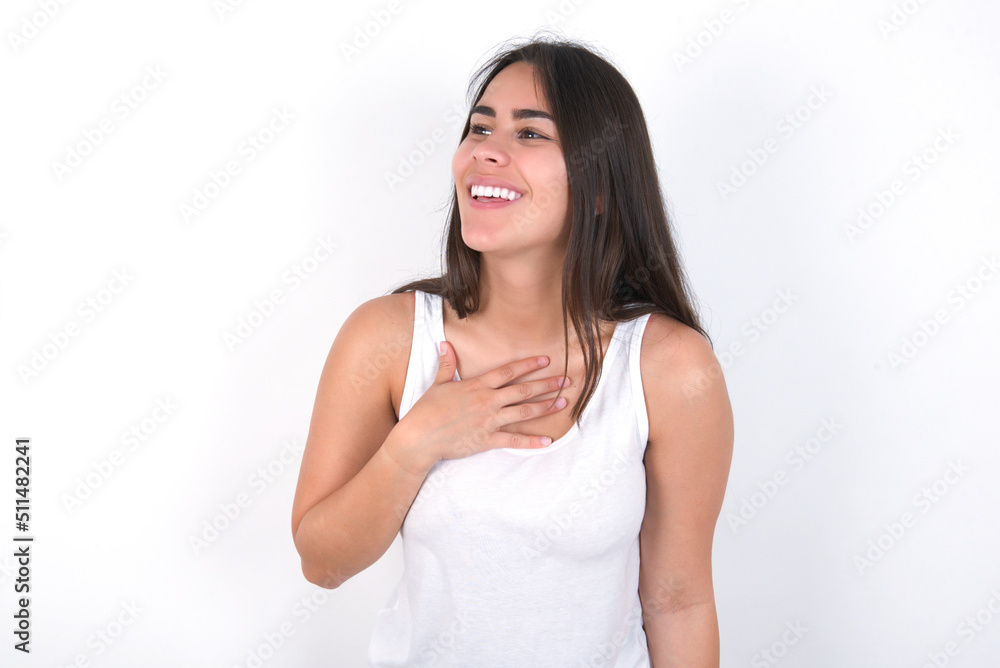 Joyful young beautiful brunette woman wearing white top over white wall expresses positive emotions recalls something funny keeps hand on chest and giggles happily. Joyful young beautiful brunette woman wearing white top over white wall expresses positive emotions recalls something funny keeps hand on chest and giggles happily.