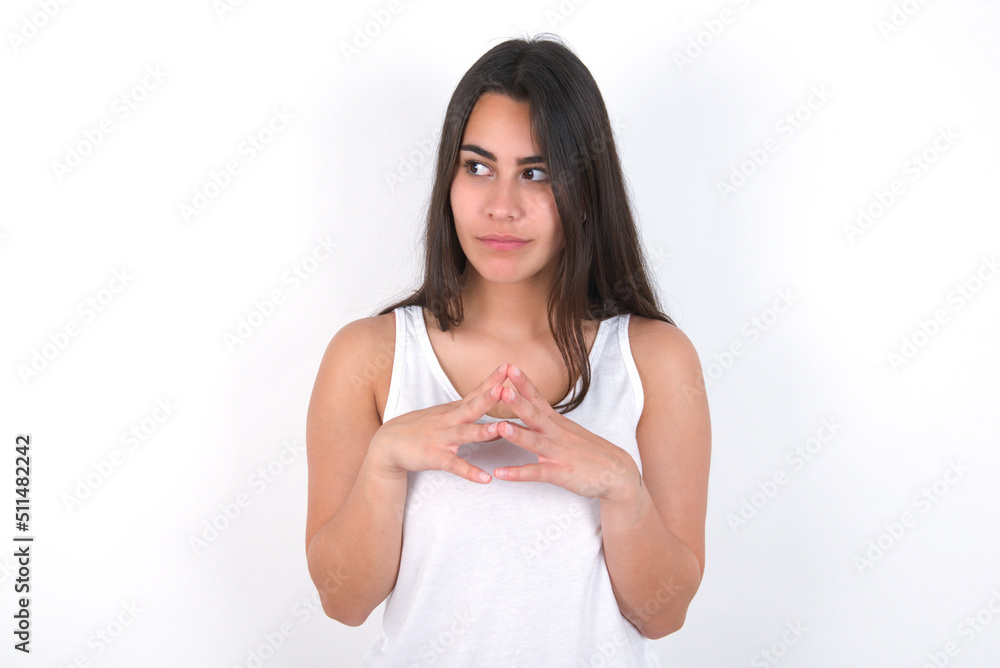 young beautiful brunette woman wearing white top over white wall steepled fingers and looks mysterious aside has great evil plan in mind young beautiful brunette woman wearing white top over white wall steepled fingers and looks mysterious aside has great evil plan in mind