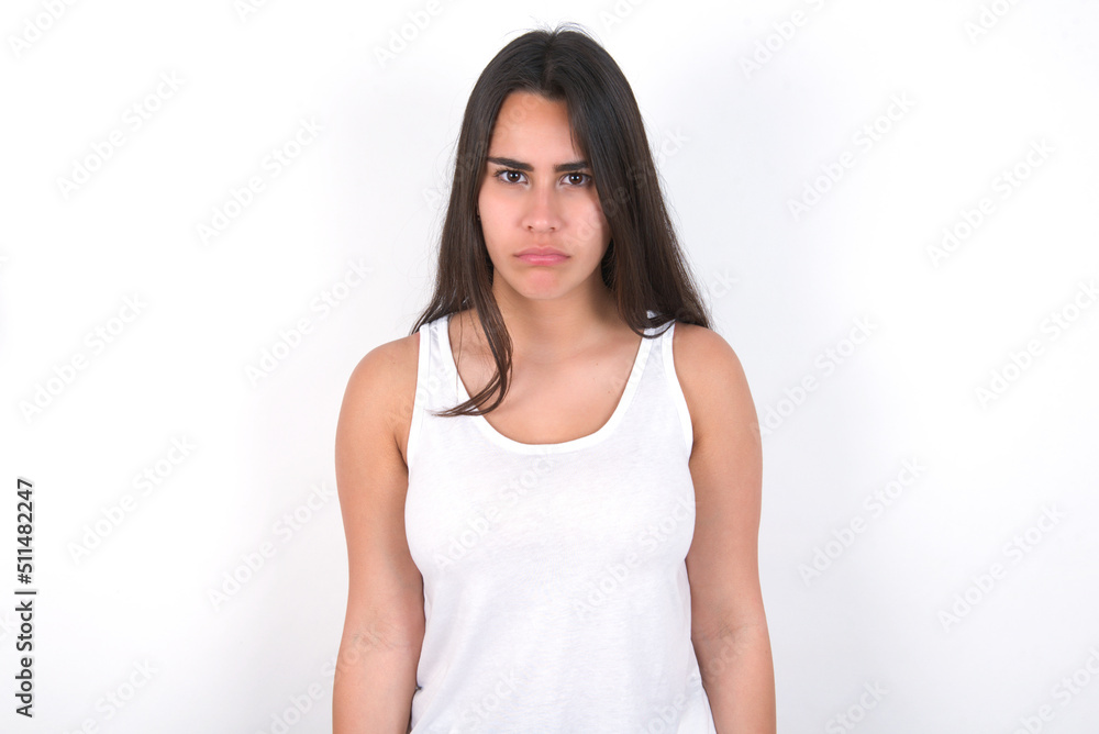 Offended dissatisfied young beautiful brunette woman wearing white top over white wall with moody displeased expression at camera being disappointed by something Offended dissatisfied young beautiful brunette woman wearing white top over white wall with moody displeased expression at camera being disappointed by something