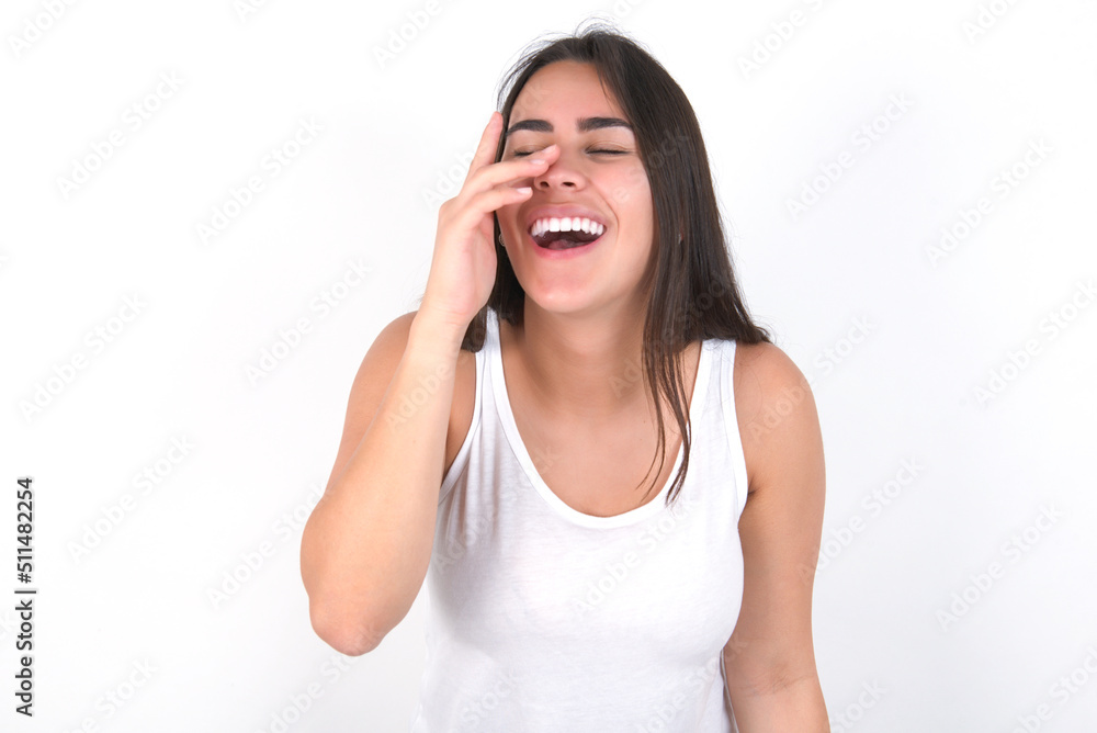 young beautiful brunette woman wearing white top over white wall makes face palm and smiles broadly, giggles positively hears funny joke poses young beautiful brunette woman wearing white top over white wall makes face palm and smiles broadly, giggles positively hears funny joke poses