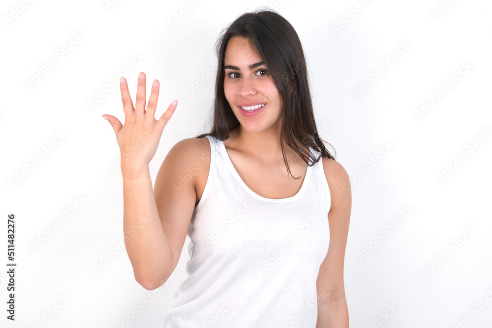 young beautiful brunette woman wearing white top over white wall smiling and looking friendly, showing number five or fifth with hand forward, counting down young beautiful brunette woman wearing white top over white wall smiling and looking friendly, showing number five or fifth with hand forward, counting down