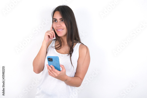 Image of a thinking dreaming Young beautiful brunette woman wearing white top over white wall using mobile phone and holding hand on face. Taking decisions and social media concept.