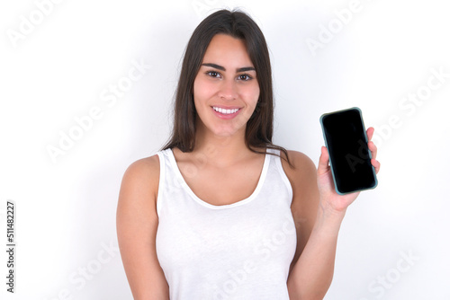 Smiling Young beautiful brunette woman wearing white top over white wall showing empty phone screen. Advertisement and communication concept.